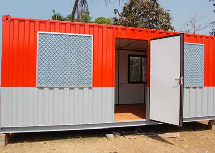 A red and grey porta cabin, designed for use as an office, stands on a sunny day. The cabin has two windows with grates and an open white door revealing a simple interior. This image could represent one of the top 10 porta cabins for offices in Jeddah.