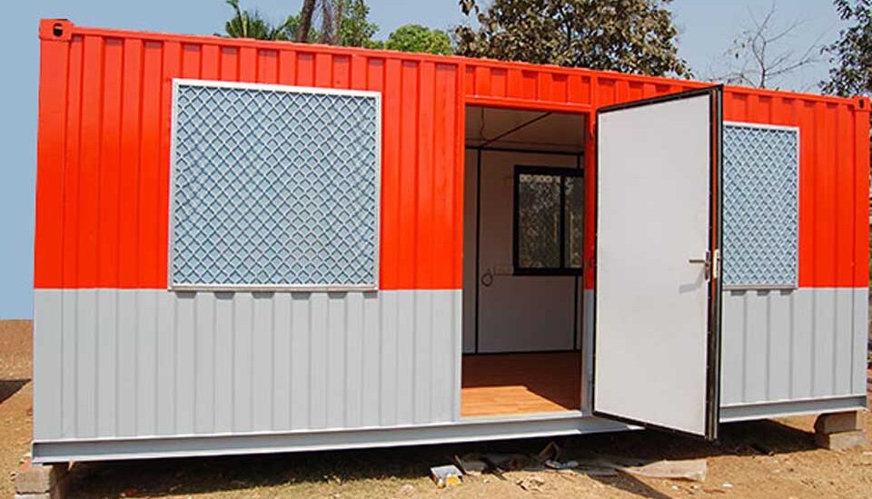 A red and grey porta cabin, designed for use as an office, stands on a sunny day. The cabin has two windows with grates and an open white door revealing a simple interior. This image could represent one of the top 10 porta cabins for offices in Jeddah.